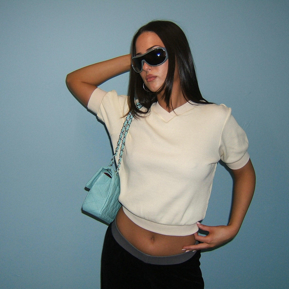 A woman posing in a cream short-sleeve knit polo crop top, black pants, black sunglasses, and carrying a light blue quilted shoulder bag against a solid blue background.