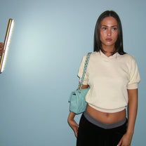 A woman stands against a light blue wall wearing a cream short-sleeve cropped top, black pants, and a light blue quilted shoulder bag with a chain strap. A hand holding a vertical light panel is visible on the left side of the frame.