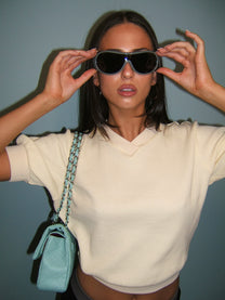 A woman wearing oversized dark sunglasses with a gray frame, a cream ribbed knit top, and a light blue quilted shoulder bag, posing against a muted blue-gray wall.