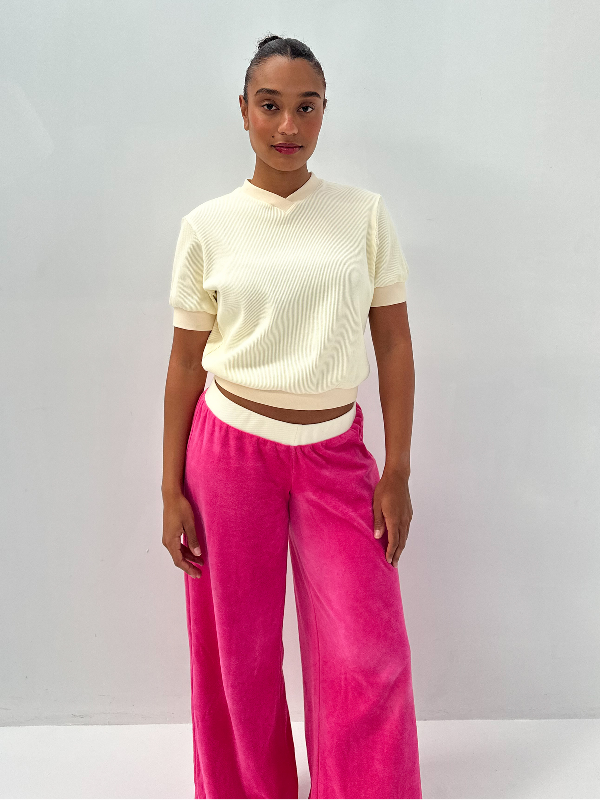 A model stands against a light gray studio background wearing a cream short-sleeve cropped knit top and bright pink wide-leg velvet pants.