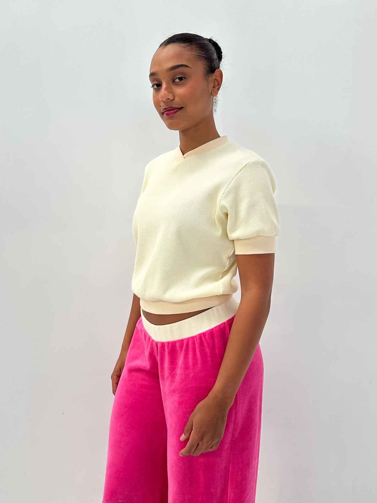 A model wearing a cream short-sleeve cropped knit top and bright pink wide-leg velvet pants, standing against a plain light gray studio background.